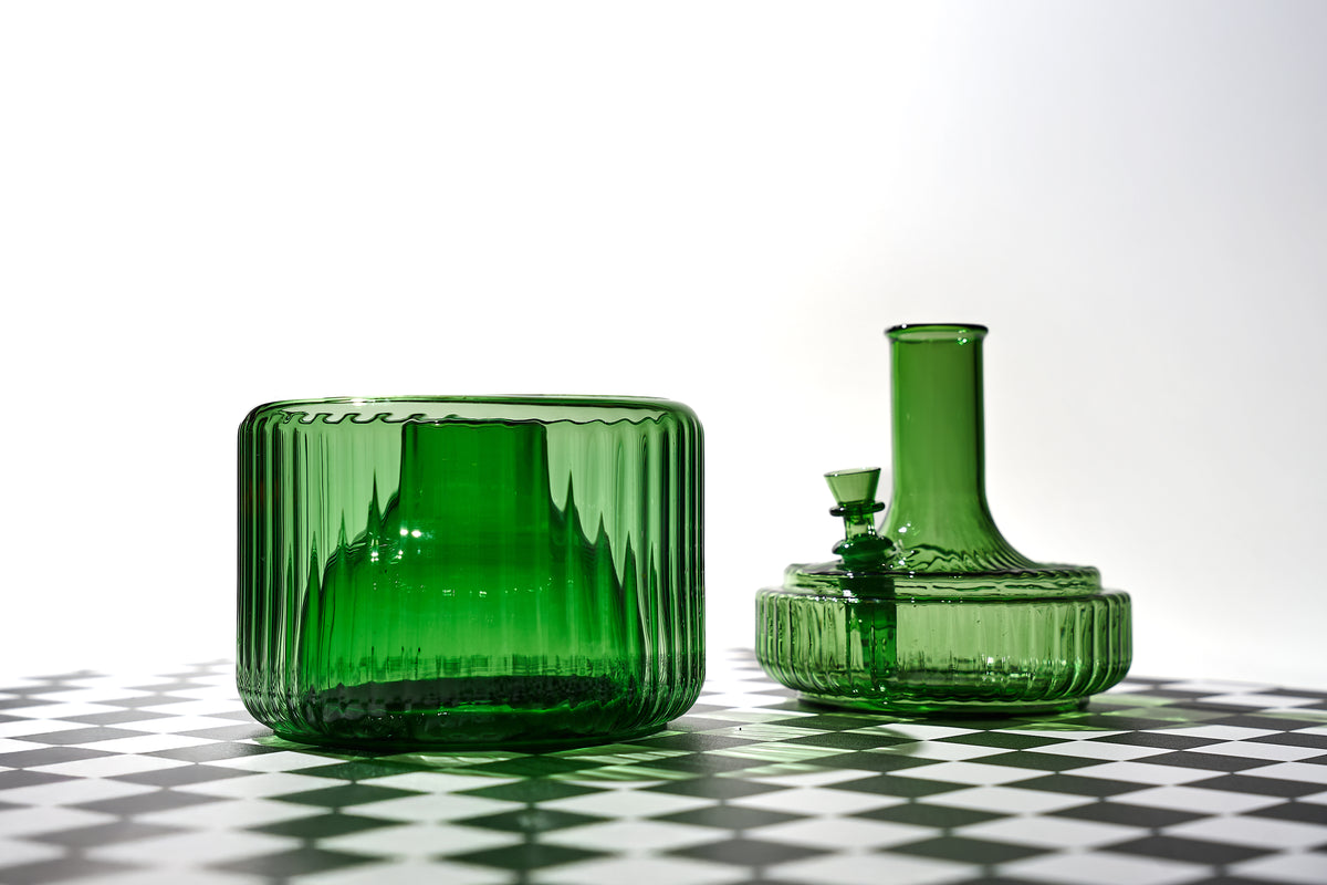 The Vase by Mantelpiece in green glass, shown in its two separate components—a sculptural flower vase and a discreet water pipe—displayed on a black-and-white checkered surface with soft lighting.