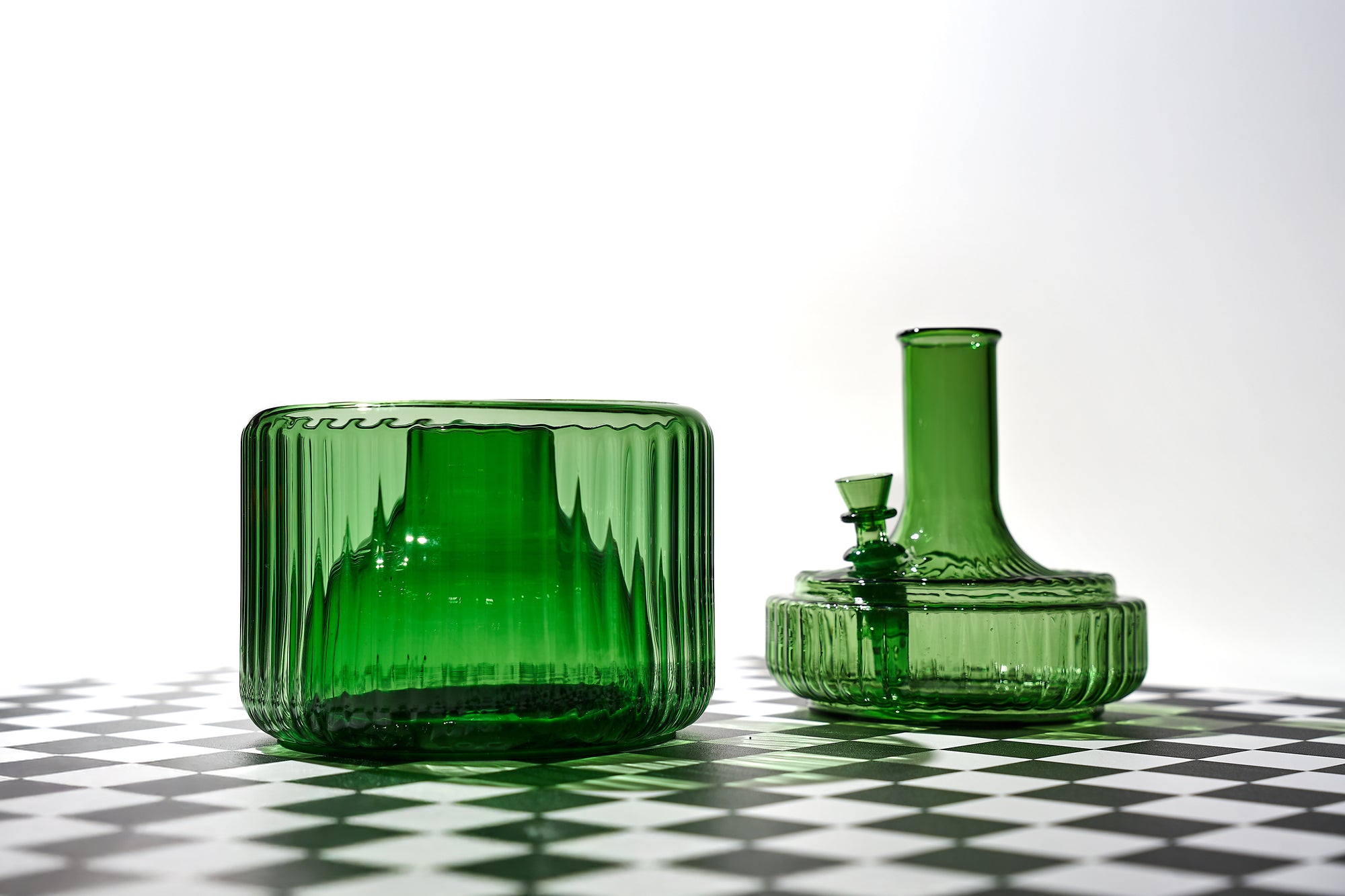 The Vase by Mantelpiece in green shown disassembled into its two parts: a sculptural green glass base and a removable top with bowl insert. Photographed on a bold checkered surface, this elegant water pipe doubles as a decorative object and bud vase alternative.