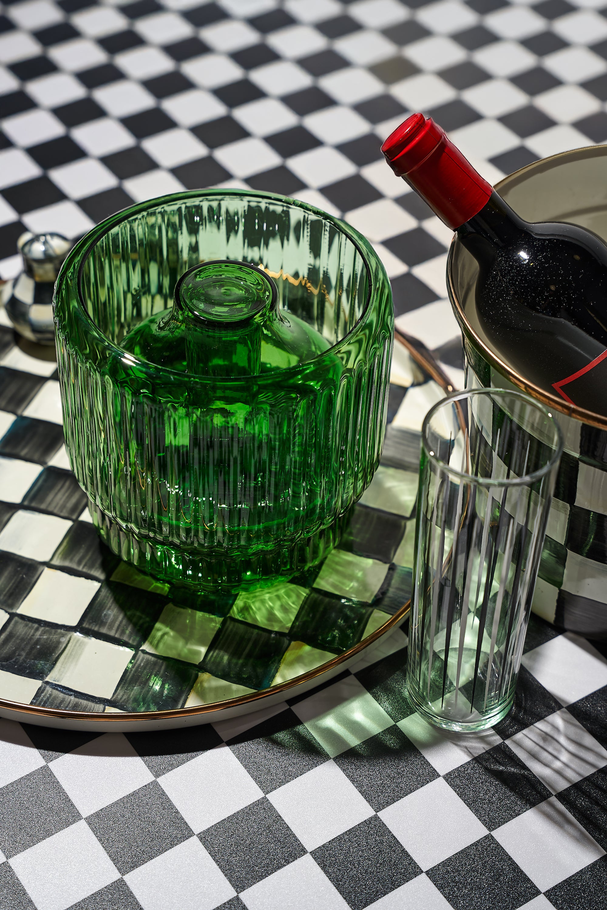 Green ribbed vase bong styled on a black and white checkered tray with a wine bottle, tall glassware, and gold accents. A refined cannabis accessory that blends seamlessly into entertaining setups, offering an elevated alternative to typical bud vases.