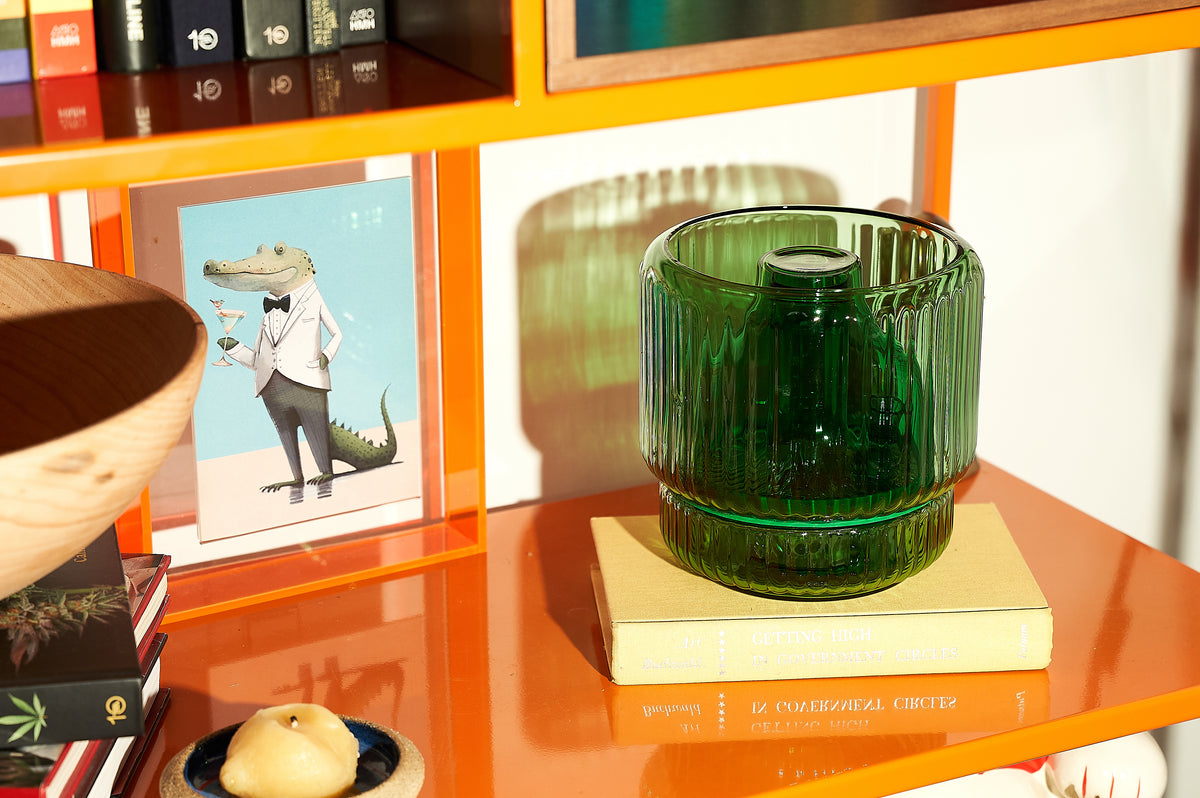 A green ribbed glass vase bong styled discreetly on a modern orange bookshelf, sitting atop a book titled Getting High in Government Circles. A clever and decorative bud vase alternative that blends into playful home decor with a mid-century vibe.
