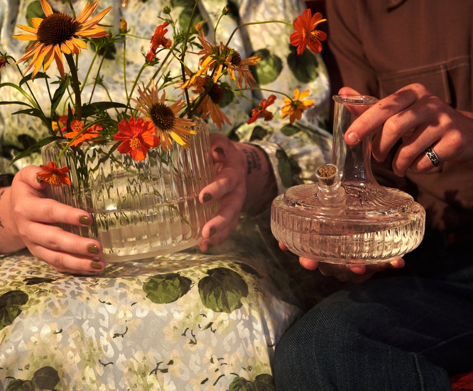 Two people sitting side by side, one holding a glass vase filled with wildflowers and the other holding a glass bong. Both pieces have a similar design, and the scene showcases a relaxed moment with natural, floral elements and casual interaction.