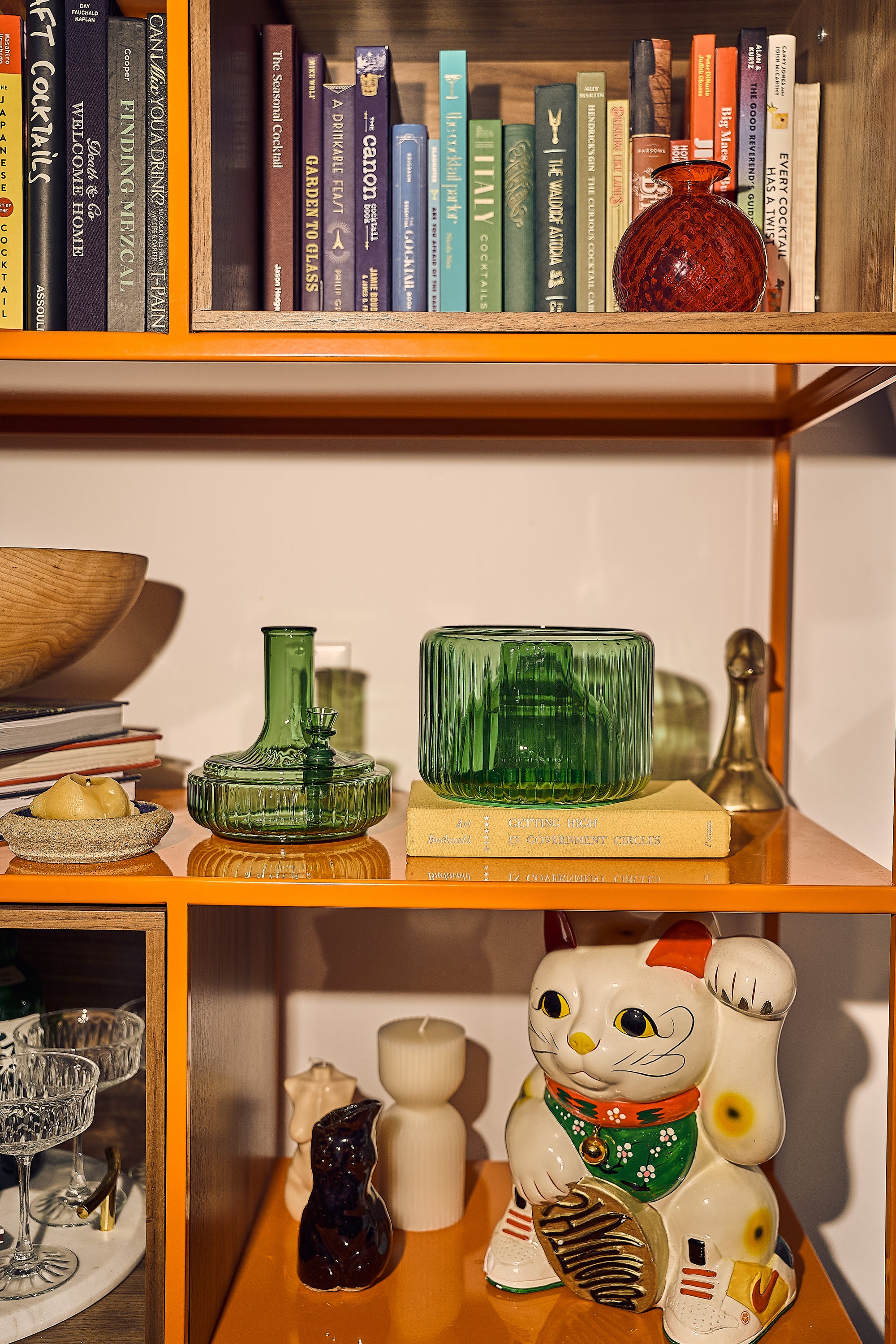 Eclectic home shelving display featuring two green glass vases—including a modular vase bong—surrounded by vintage books, a maneki-neko cat, and barware. A curated cannabis-friendly shelf styled to look like a high-design bud vase setup.