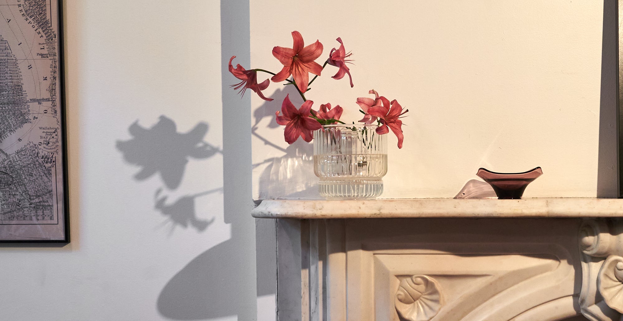 A clear fluted glass vase bong styled as a flower arrangement with pink lilies, placed on a white marble mantel with soft shadows cast against the wall. This modern cannabis accessory is designed to look like a bud vase, offering a discreet and decorative bong option that fits into any sophisticated living space. Ideal for those looking for a stylish smoking piece that doesn’t sacrifice design.