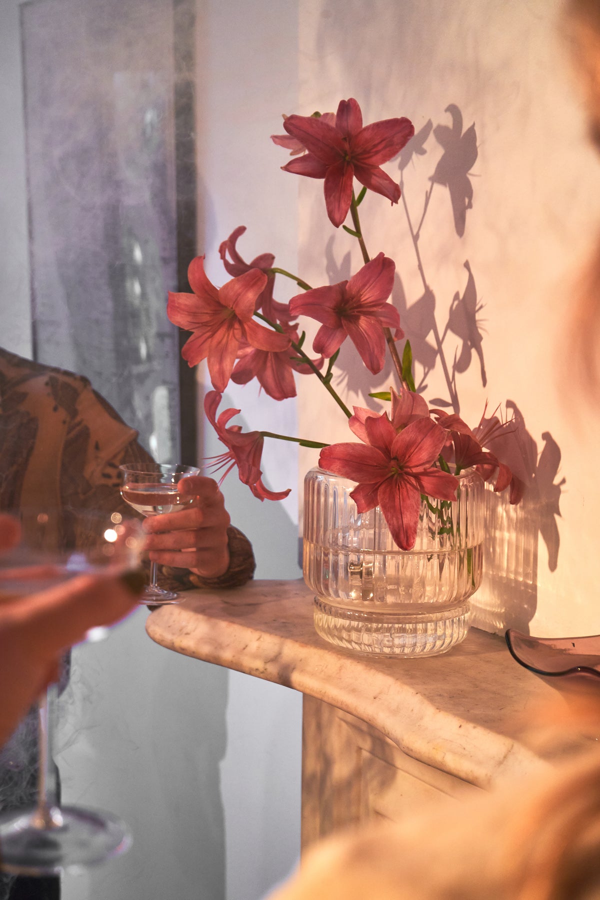 A group gathers near a white marble mantel, where a clear ribbed glass bong vase is repurposed as a floral centerpiece holding pink lilies. The scene is softly lit, evoking a sense of relaxed elegance. This beautiful bong blends into the decor, functioning as both a flower vase and a discreet smoking piece—perfect for cannabis users who value design-forward, home-friendly accessories.