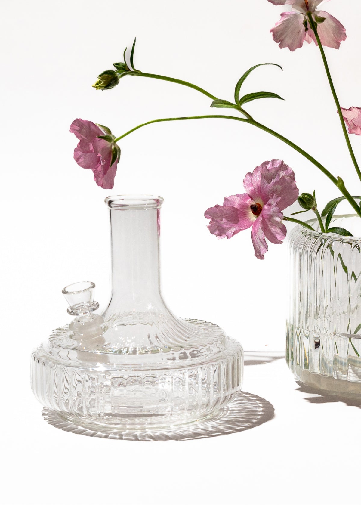 Clear glass vase bong base beside ribbed flower vase with pink blooms on white background