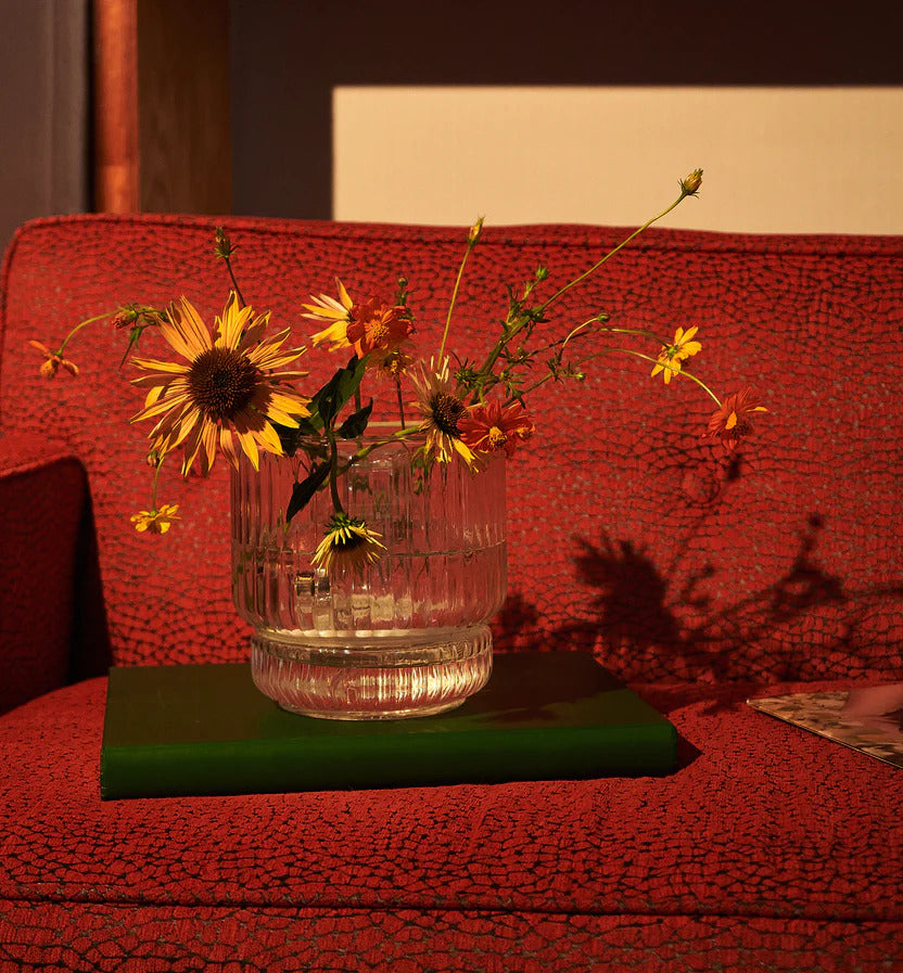 A clear ribbed glass vase bong used as a flower vase, resting on a green book, on a deep red textured couch. Filled with fresh yellow and orange wildflowers, this discreet bong doubles as a decorative object, seamlessly blending into eclectic home decor. An elegant and functional alternative to traditional bongs, perfect for those searching for a beautiful, aesthetic bong.