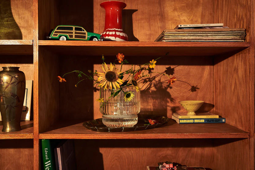 A clear ribbed glass bong vase sits on a mid-century wood bookshelf, filled with sunflowers and wildflowers, catching warm afternoon light. Nestled between vintage books and ceramics, this discreet bong is styled as a flower vase, offering a clever solution for anyone seeking a beautiful bong that doesn’t disrupt their home aesthetic.