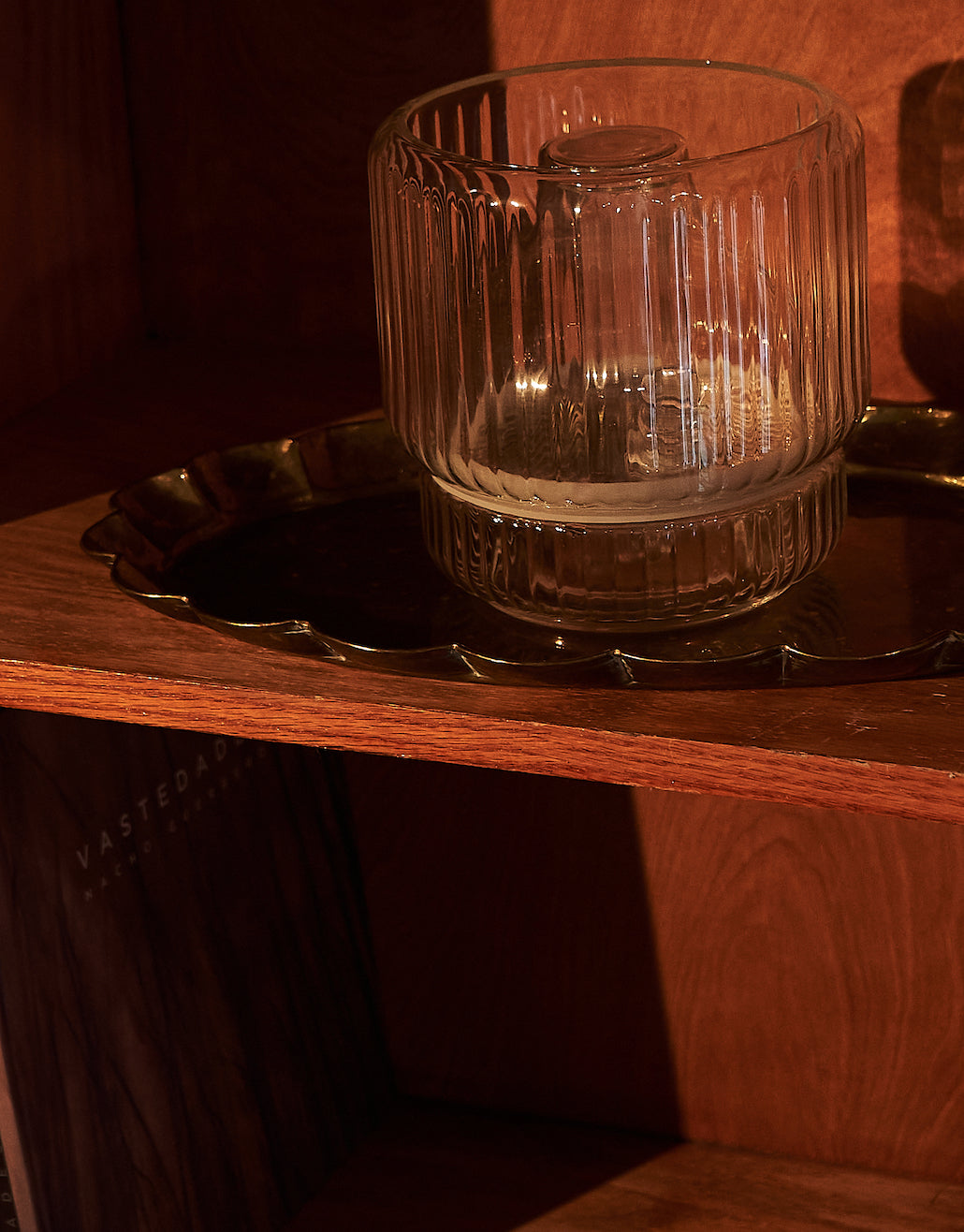 A clear glass vase placed on a brass tray, sitting on a wooden shelf. The warm lighting highlights the vase's ridged texture, creating soft reflections on the surface beneath it.