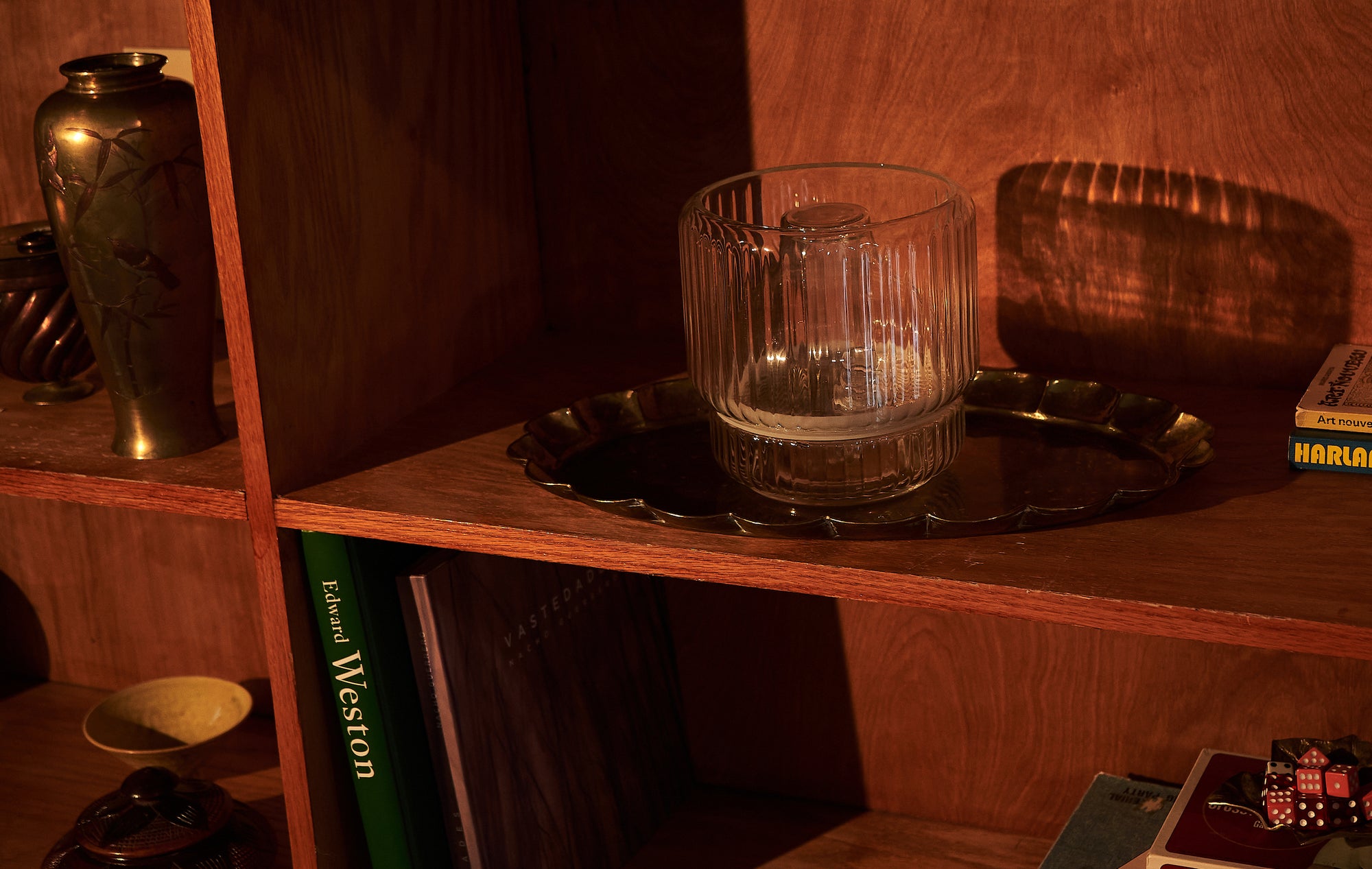 A clear glass vase placed on a brass tray, sitting on a wooden shelf. The warm lighting highlights the vase's ridged texture, creating soft reflections on the surface beneath it.