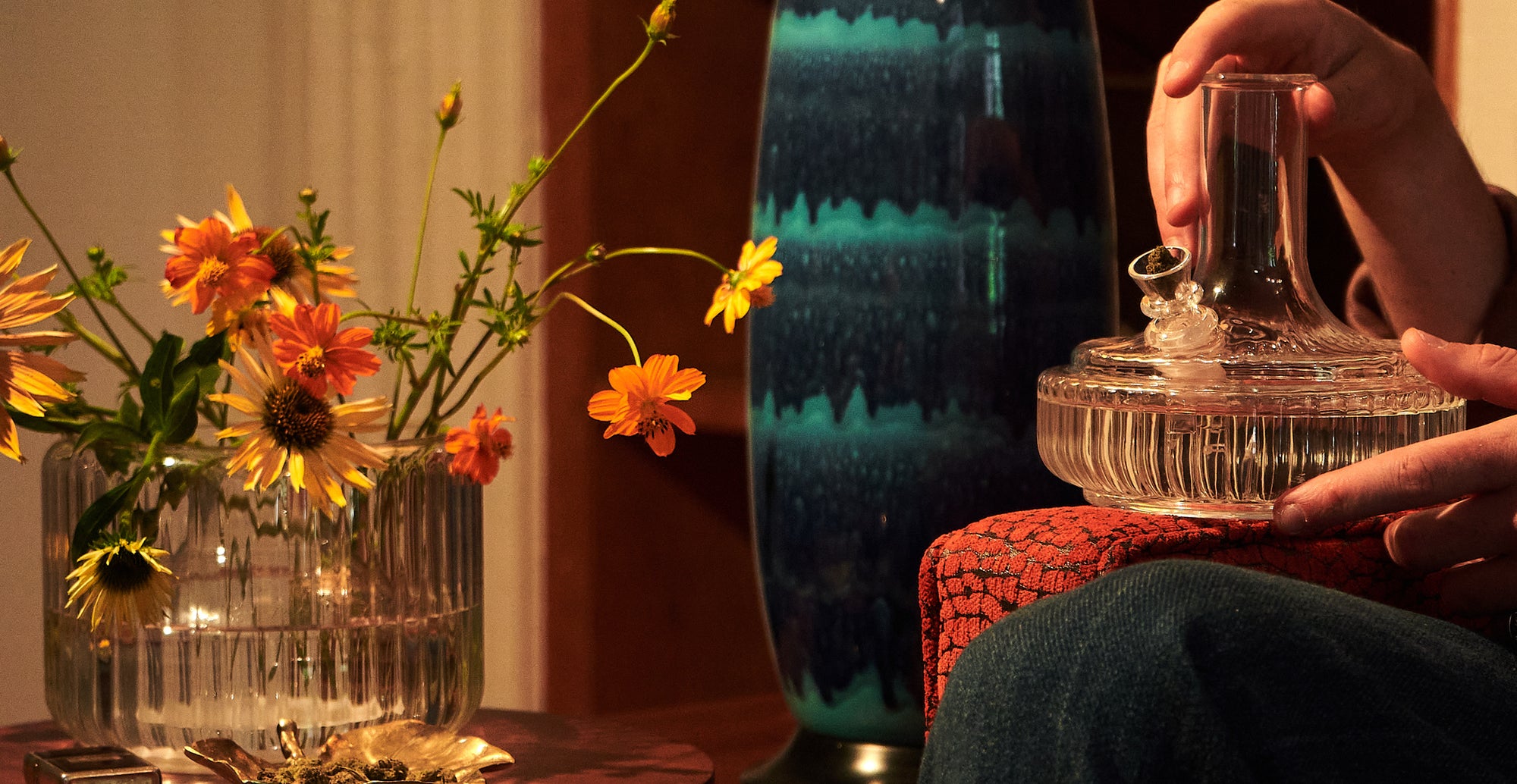 This stylish scene features Mantelpiece's innovative bong that doubles as a vase, perfectly blending functionality and home decor. On the left, a clear glass vase filled with vibrant orange flowers adds a pop of color, while on the right, a hand carefully holds the Mantelpiece bong—a piece that seamlessly integrates into any home setting. Designed with both form and function in mind, this elegant glass bong is a hidden gem, perfect for those who appreciate high-end, vintage-inspired home decor. Discover how