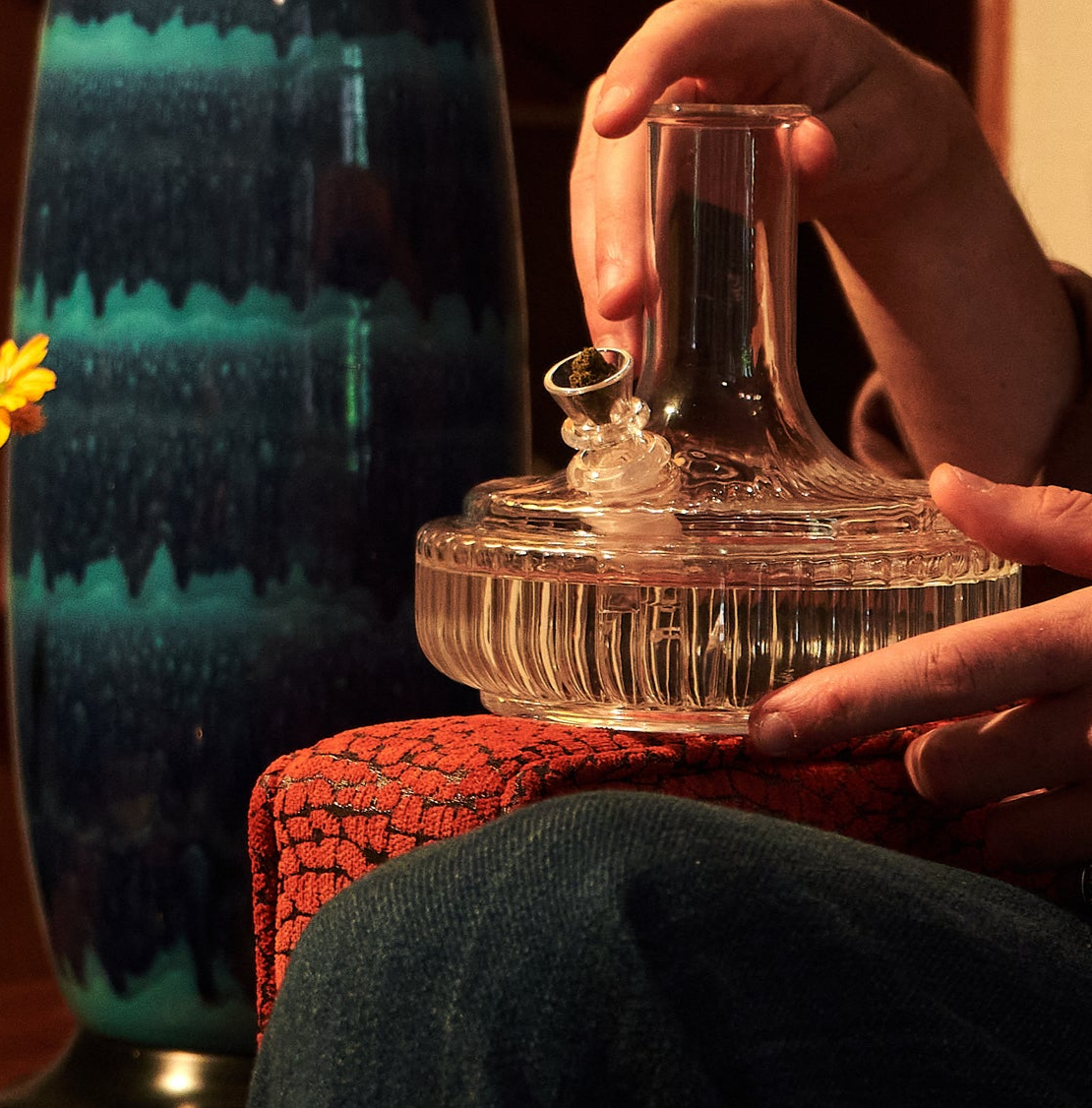 A person holds a clear glass bong, with their hand resting on a red textured couch arm. The intricate ridged design of the bong reflects the warm lighting, while a blue mid-century modern vase is partially visible in the background, adding a retro touch to the scene.