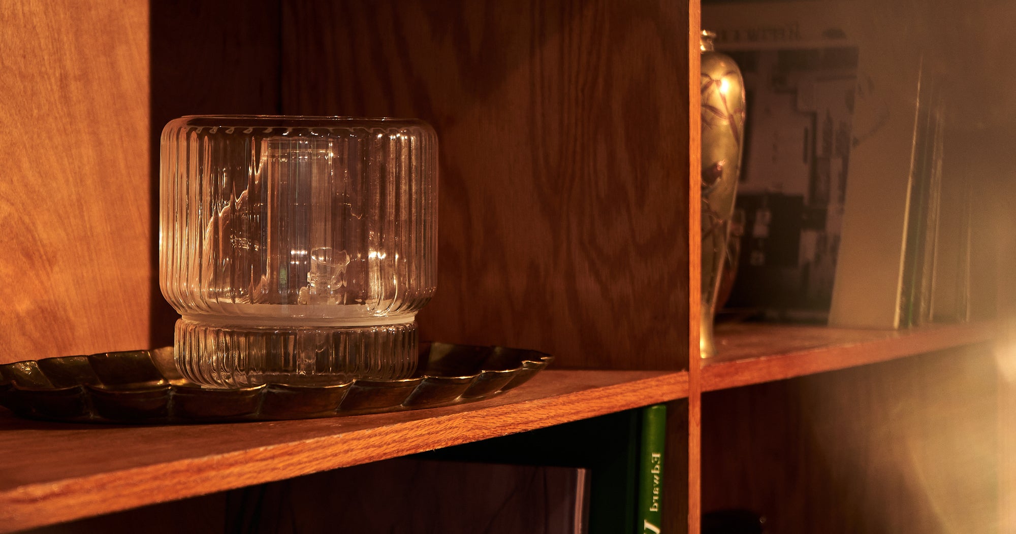 A beautifully lit Mantelpiece bong vase, crafted from clear ribbed glass, sits atop a dark brass tray on a wooden shelf. The warm wood tones and subtle reflections of light give the scene a cozy, vintage feel. The Mantelpiece vase blends seamlessly with the decor, evoking both elegance and discretion in a stylish living space.