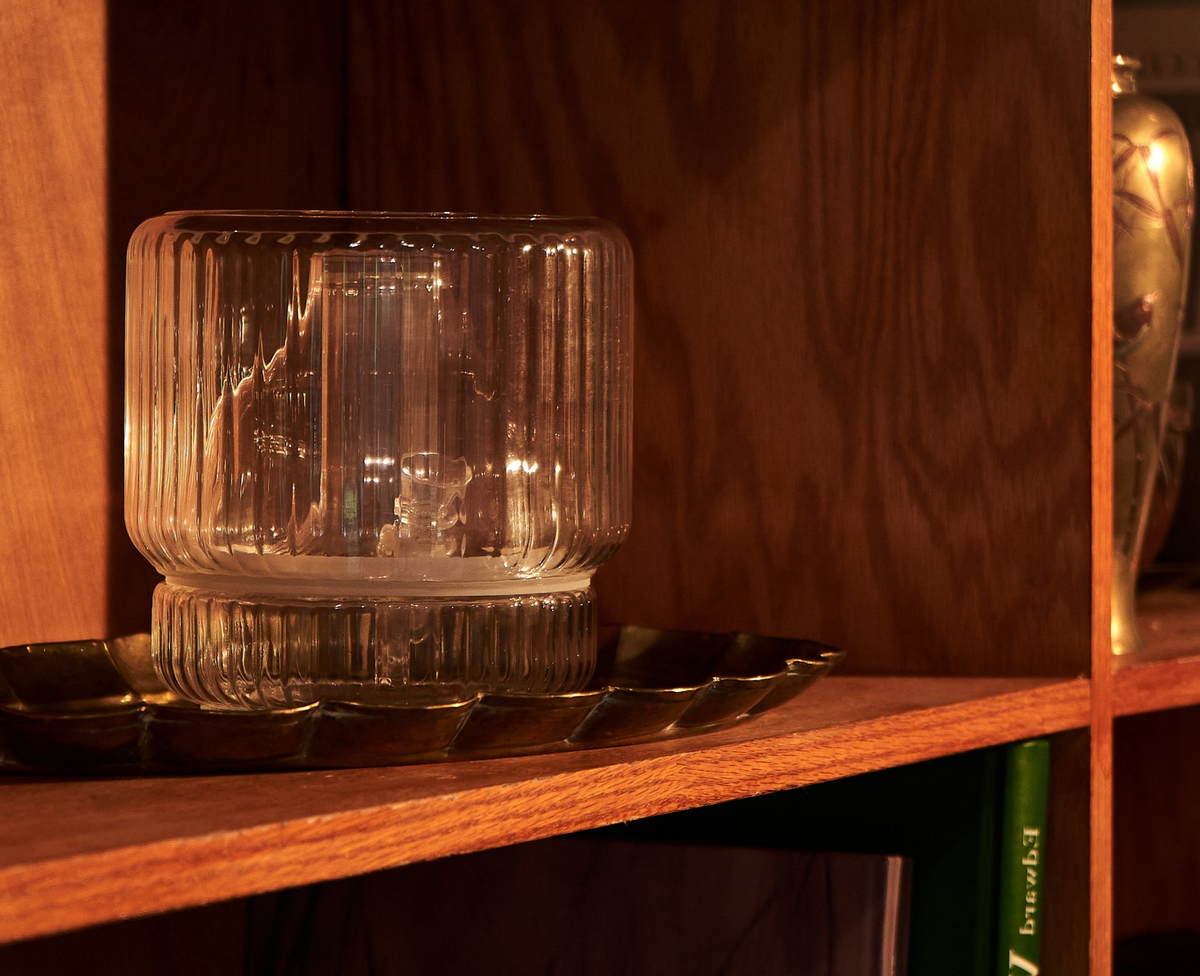 A clear glass vase sits on a brass tray on a wooden mid-century modern bookshelf. The warm light reflects off the vase's ridged surface, casting subtle shadows. In the background, a brass vase and books add to the carefully curated vintage decor.